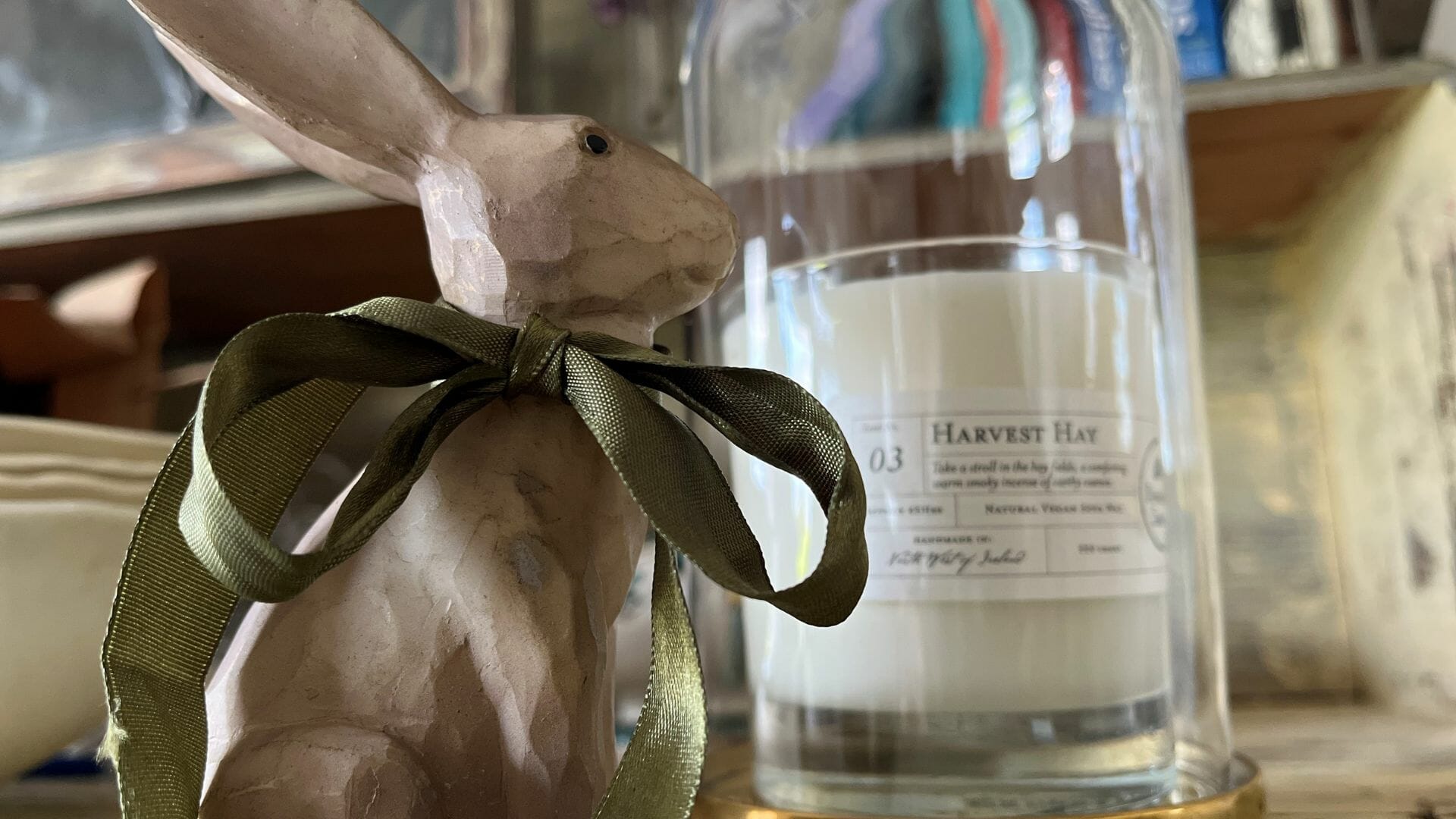 Wooden hare with green bow looking at white candle in glass dome.