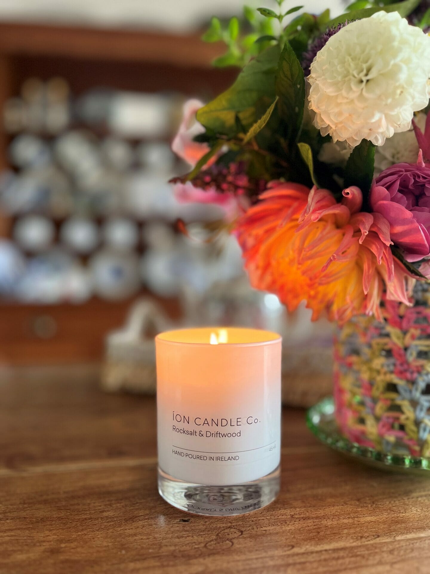 Scented candle, Ion, in white glass, sitting on table beside flowers